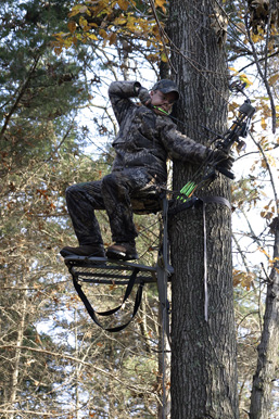 Un chasseur à l'arc en habit de camouflage se tient prêt à décocher du haut de sa plateforme, dans un arbre.