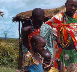 Two African adults and two young African children wearing colorful clothes are posing near a hut without looking at the camera. 